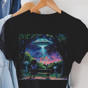 May include: A black t-shirt with a colorful graphic print of a flying saucer hovering over a forest scene. Two blue alien figures stand near a woman sitting on a bench. The sky is filled with stars and a pink and purple glow.