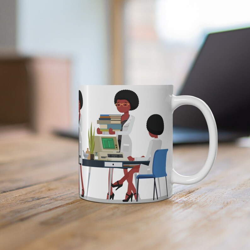 K&ouml;nnte beinhalten: Wei&szlig;e Keramiktasse mit einer bunten Illustration von zwei Frauen, die an einem Schreibtisch mit einem Computer und B&uuml;chern arbeiten. Die Frauen tragen wei&szlig;e Laborkittel und haben dunkle Haut und schwarze Haare.