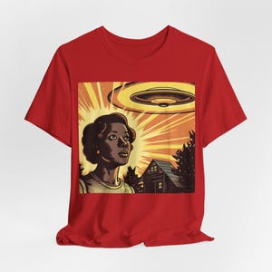 May include: A red t-shirt with a graphic print of a woman looking up at a flying saucer. The graphic is in a retro style with a yellow, orange, and brown color scheme. The woman is wearing a light blue shirt and has dark hair. The flying saucer is yellow and has a brown underside. There is a small brown house in the background.