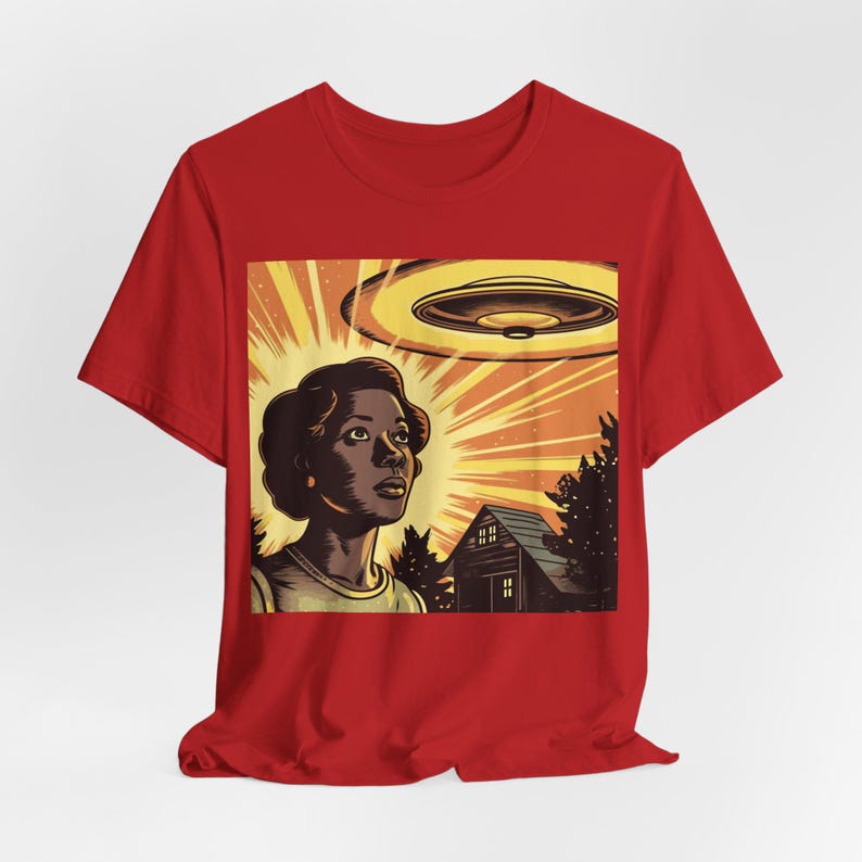 May include: A red t-shirt with a graphic print of a woman looking up at a flying saucer. The graphic is in a retro style with a yellow, orange, and brown color scheme. The woman is wearing a light blue shirt and has dark hair. The flying saucer is yellow and has a brown underside. There is a small brown house in the background.