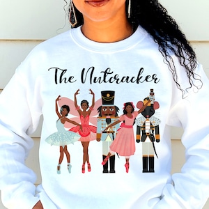 Könnte beinhalten: Weißer Pullover mit dem Text "The Nutcracker" in Schwarz gedruckt. Das Design zeigt eine vielfältige Gruppe von Figuren aus dem Ballett, darunter ein Nussknacker, ein Mäusekönig und Ballerinas.
