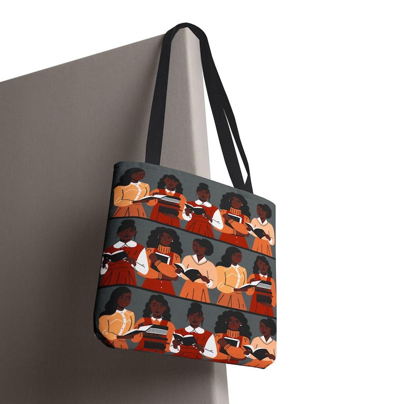 Puede incluir: Un bolso tote con un patr&oacute;n repetido de mujeres leyendo libros. Las mujeres est&aacute;n representadas con varios atuendos, incluyendo vestidos naranjas, rojos y amarillos. El bolso tiene asas negras y un fondo gris.