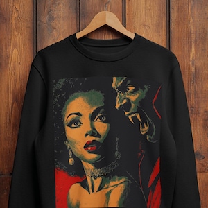 May include: Black sweatshirt featuring a vintage-style illustration of a woman and a vampire. The woman has red lips and is wearing a necklace and earrings. The vampire has green skin and red accents. The design is set against a red background.