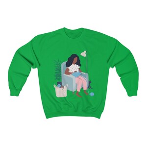 May include: A green sweatshirt with a graphic of a person sitting in a chair knitting with a cat sitting next to them.