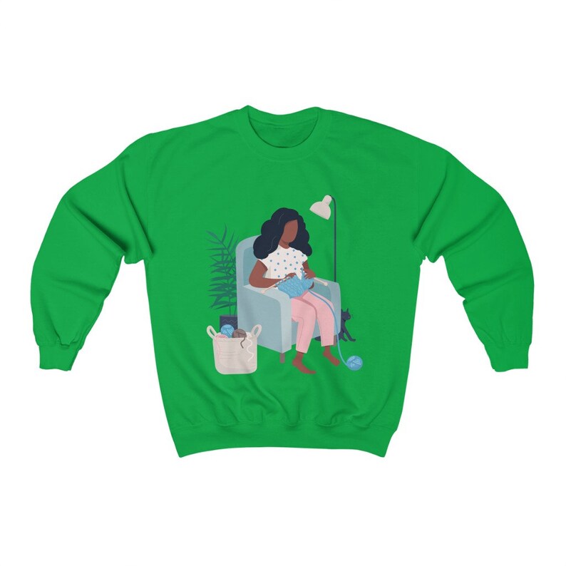 May include: A green sweatshirt with a graphic of a person sitting in a chair knitting with a cat sitting next to them.