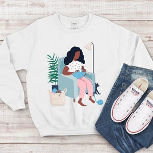 May include: A white sweatshirt with a graphic of a person sitting in a chair knitting. The person has dark hair and is wearing a pink dress with white polka dots. There is a cat sitting on the floor next to the chair. The sweatshirt is on a white wooden surface with a pair of blue jeans and white sneakers.