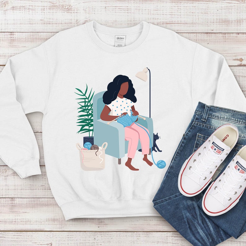 May include: A white sweatshirt with a graphic of a person sitting in a chair knitting. The person has dark hair and is wearing a pink dress with white polka dots. There is a cat sitting on the floor next to the chair. The sweatshirt is on a white wooden surface with a pair of blue jeans and white sneakers.