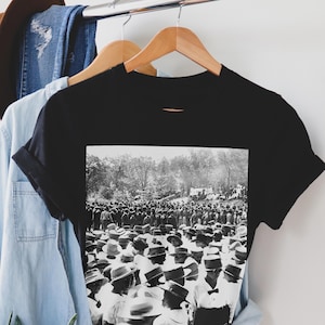 Puede incluir: Camiseta negra con una imagen en blanco y negro de una multitud de personas con sombreros, reunidas al aire libre. La imagen est&aacute; impresa en la parte delantera de la camiseta, con las mangas enrolladas. La camiseta cuelga de una percha de madera.