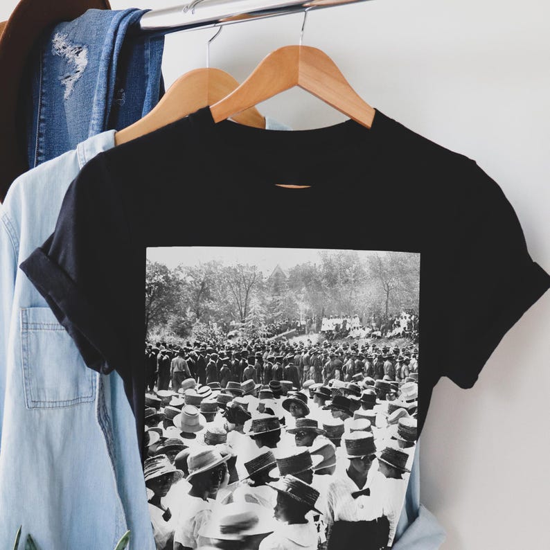 Puede incluir: Camiseta negra con una imagen en blanco y negro de una multitud de personas con sombreros, reunidas al aire libre. La imagen est&aacute; impresa en la parte delantera de la camiseta, con las mangas enrolladas. La camiseta cuelga de una percha de madera.