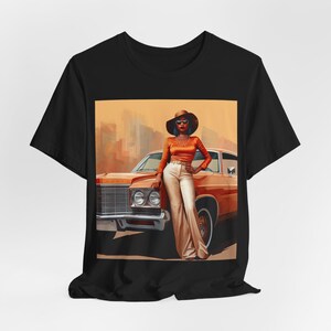 Op de afbeelding: Een zwart T-shirt met een grafische print van een vrouw in een bruine hoed en een oranje outfit, die naast een oranje oldtimer staat.