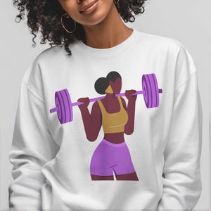 May include: White sweatshirt with a graphic of a woman lifting a barbell. The woman is in purple, yellow, and brown. The barbell and weights are purple, and the woman is wearing a yellow top and purple shorts. A fitness-themed design.
