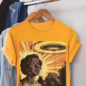 May include: A yellow t-shirt with a graphic print of a woman looking up at a flying saucer. The graphic is in a retro comic book style with a bright orange, yellow, and red color scheme.