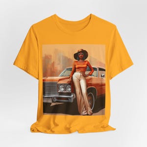 Op de afbeelding: Een geel T-shirt met een grafische print van een vrouw in een bruine hoed en zonnebril die naast een vintage auto staat. De vrouw draagt een bruin topje, een witte broek en een ketting. De auto is bruin en heeft een wit interieur.