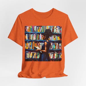 Może przedstawiać: Pomarańczowy T-shirt z grafiką przedstawiającą osobę stojącą przed regałem wypełnionym kolorowymi książkami. Osoba trzyma książkę i patrzy na nią.