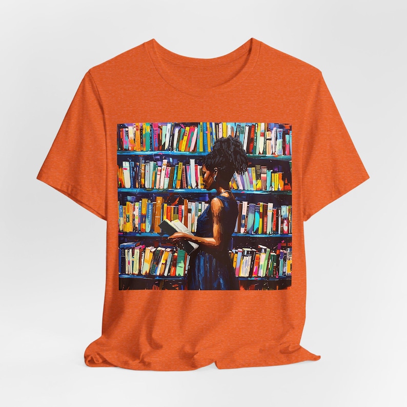 Może przedstawiać: Pomarańczowy T-shirt z grafiką przedstawiającą osobę stojącą przed regałem wypełnionym kolorowymi książkami. Osoba trzyma książkę i patrzy na nią.