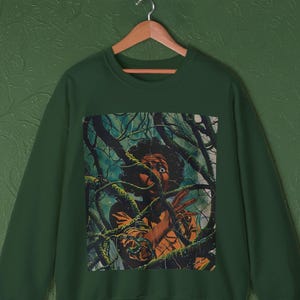 May include: Dark green sweatshirt featuring a vintage-style illustration. The graphic depicts a person with a look of fear, surrounded by dark, twisting vines. The artwork has a retro, pulp-magazine aesthetic, with a color palette of greens, browns, and blues.