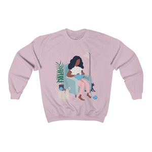 May include: A pink sweatshirt with a graphic of a person sitting in a chair knitting with a cat sitting next to them.