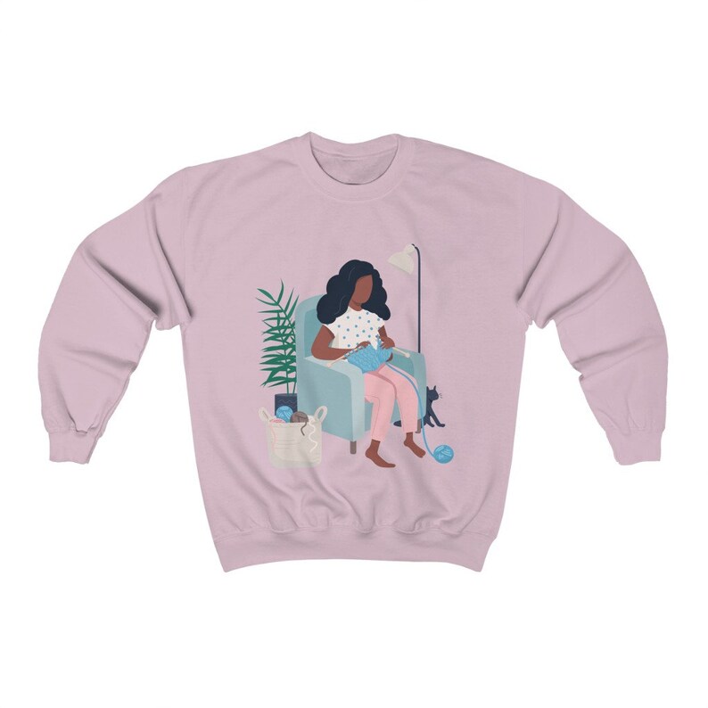 May include: A pink sweatshirt with a graphic of a person sitting in a chair knitting with a cat sitting next to them.