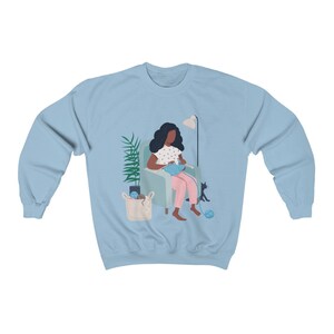 May include: A light blue sweatshirt with a graphic of a person sitting in a chair knitting with a cat sitting next to them.