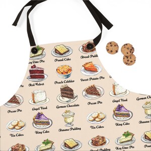 May include: A beige apron printed with illustrations of various desserts, including pies, cakes, and puddings. The apron has black ties and a button closure. Three chocolate chip biscuits are visible in the upper right corner.