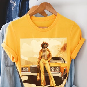 May include: A yellow t-shirt with a graphic print of a woman wearing a hat and sunglasses standing next to a yellow car.