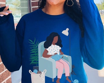 Puede incluir: Una sudadera azul con un gráfico de una mujer sentada en una silla tejiendo. La mujer lleva un vestido rosa y tiene el pelo negro. La silla es azul y tiene una lámpara blanca a su lado.