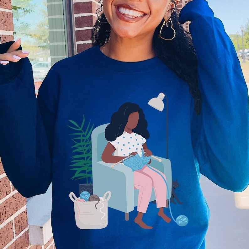 May include: A blue sweatshirt with a graphic of a woman sitting in a chair knitting. The woman is wearing a pink dress and has black hair. The chair is blue and has a white lamp next to it.