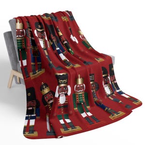 Puede incluir: Manta de forro polar roja con un patrón repetido de figuras de cascanueces negras. Los cascanueces llevan uniformes y sombreros de diferentes colores.