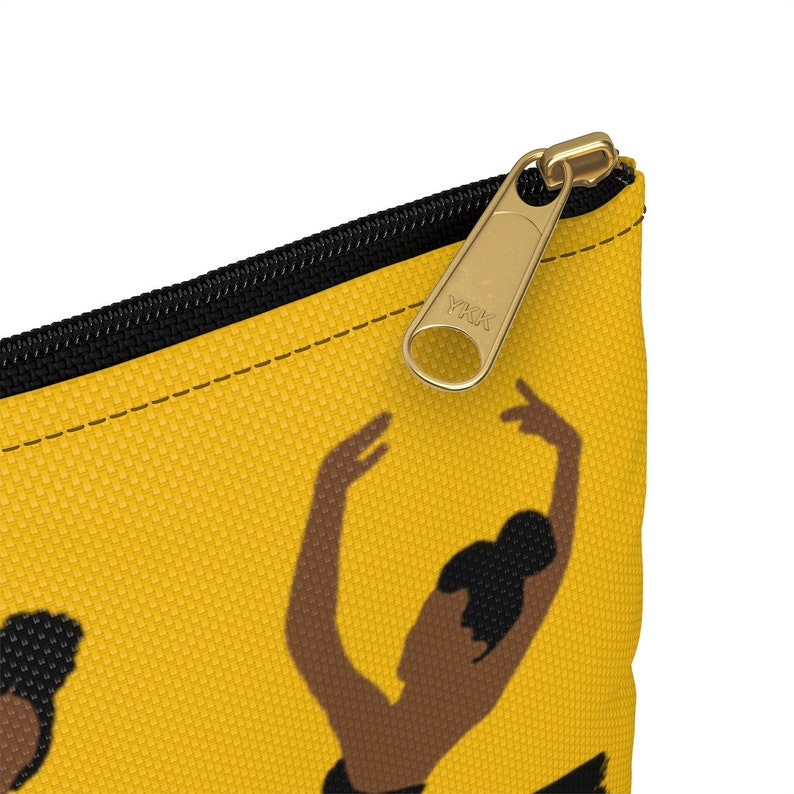 Pode incluir: Bolsa de cosm&eacute;ticos amarela mostarda com z&iacute;per preto e puxador dourado. A bolsa apresenta um padr&atilde;o repetido de uma bailarina em collant preto. O puxador do z&iacute;per tem o texto "YKK".