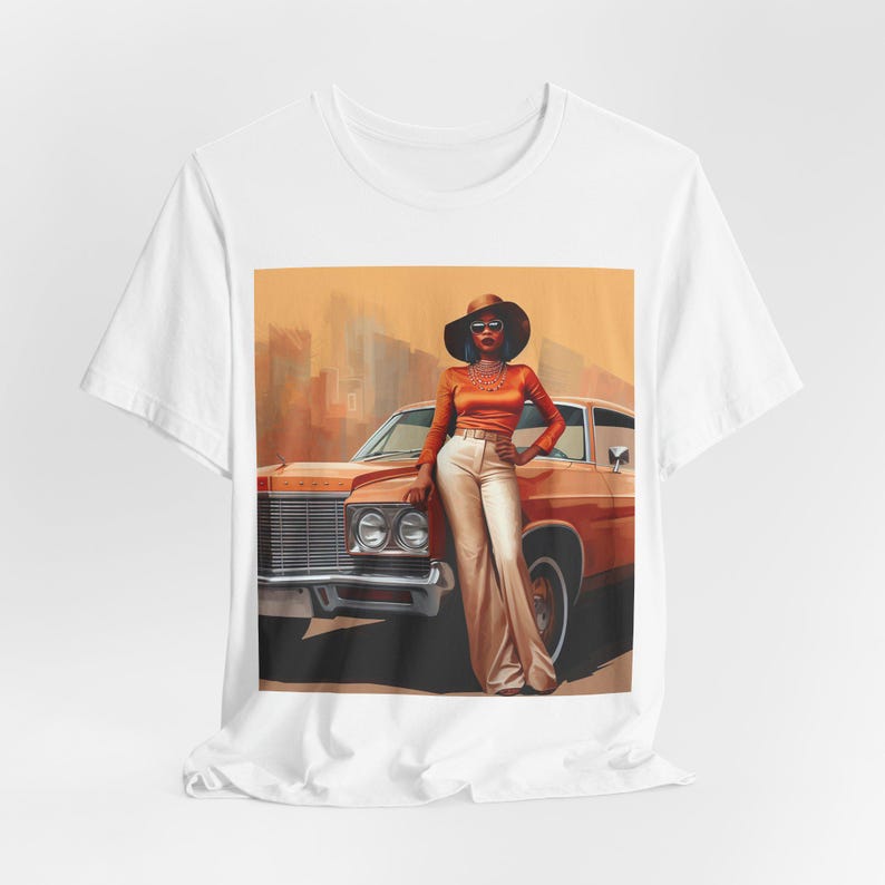 Op de afbeelding: Een wit T-shirt met een grafische print van een vrouw in een bruine hoed en een oranje outfit die naast een vintage auto staat. De achtergrond is een wazige stadssc&egrave;ne.