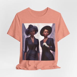 May include: A coral pink t-shirt featuring a graphic print of two women in black dresses and hats. The women are walking along a street.