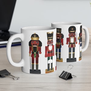 Pode incluir: Duas canecas de cerâmica branca com um tema natalino. As canecas apresentam um design de cinco figuras de quebra-nozes negros em várias poses, vestindo uniformes vermelhos, verdes e dourados. Os quebra-nozes seguram espadas, tambores e outros acessórios festivos.