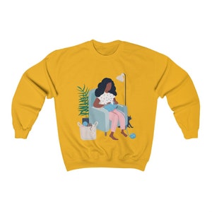 May include: A yellow sweatshirt with a graphic of a Black woman sitting in a chair knitting with a cat beside her. The woman is wearing a pink dress with white polka dots and has dark hair. The chair is blue and there is a basket of yarn on the floor.