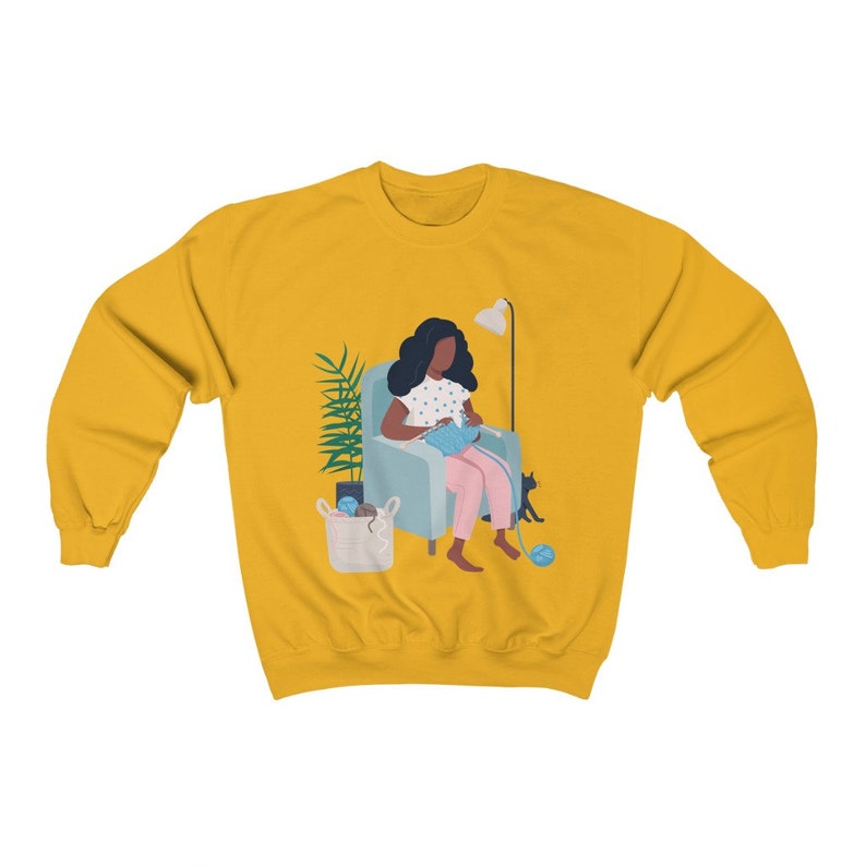 May include: A yellow sweatshirt with a graphic of a Black woman sitting in a chair knitting with a cat beside her. The woman is wearing a pink dress with white polka dots and has dark hair. The chair is blue and there is a basket of yarn on the floor.