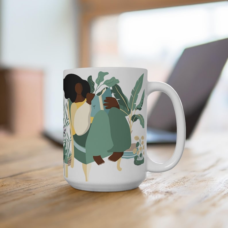 May include: White ceramic mug with a colorful illustration of a person sitting in a chair surrounded by plants. The person is holding a book and has brown skin and black hair.