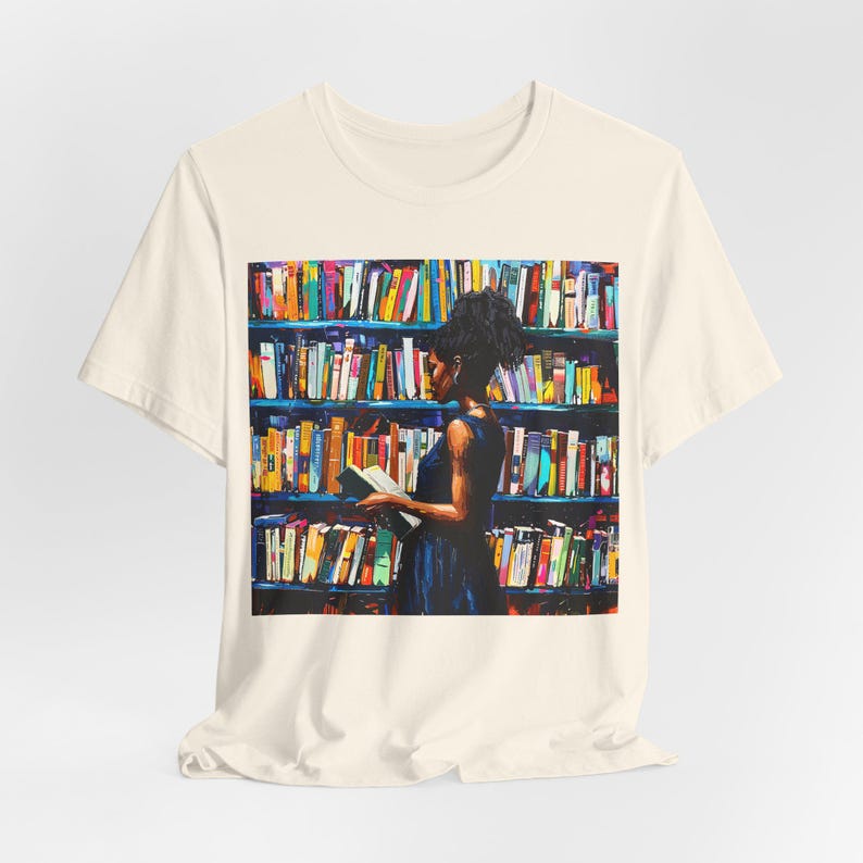 Może przedstawiać: Biały t-shirt z kolorowym nadrukiem graficznym przedstawiającym osobę stojącą przed regałem wypełnionym książkami. Osoba trzyma książkę w ręku i patrzy na nią.