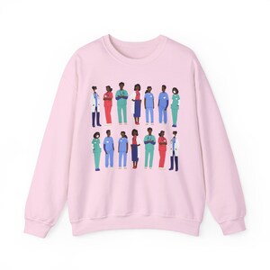 May include: Pink sweatshirt with a graphic print of a diverse group of healthcare workers wearing scrubs and masks. The graphic features a variety of skin tones and hairstyles.