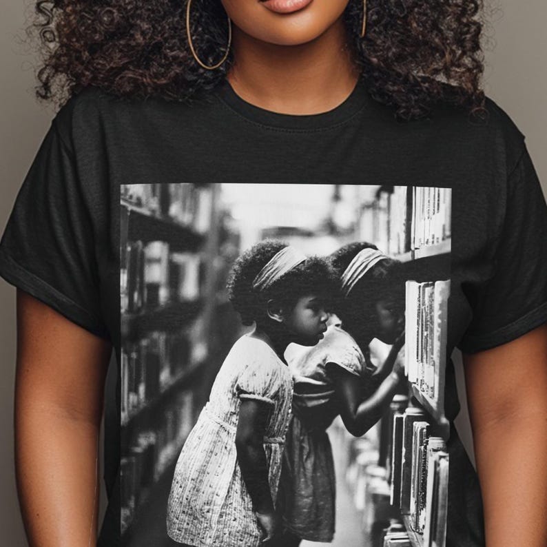 May include: Black t-shirt featuring a black and white image of two young girls in a library, examining books on a shelf. The girls are wearing dresses and headbands. The background shows bookshelves filled with books.
