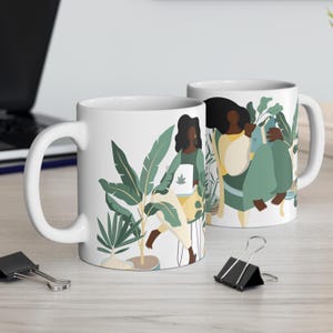 May include: Two white ceramic mugs with a design of two women standing in a jungle setting with plants and flowers. The women are wearing yellow and green clothing and have dark skin and hair.