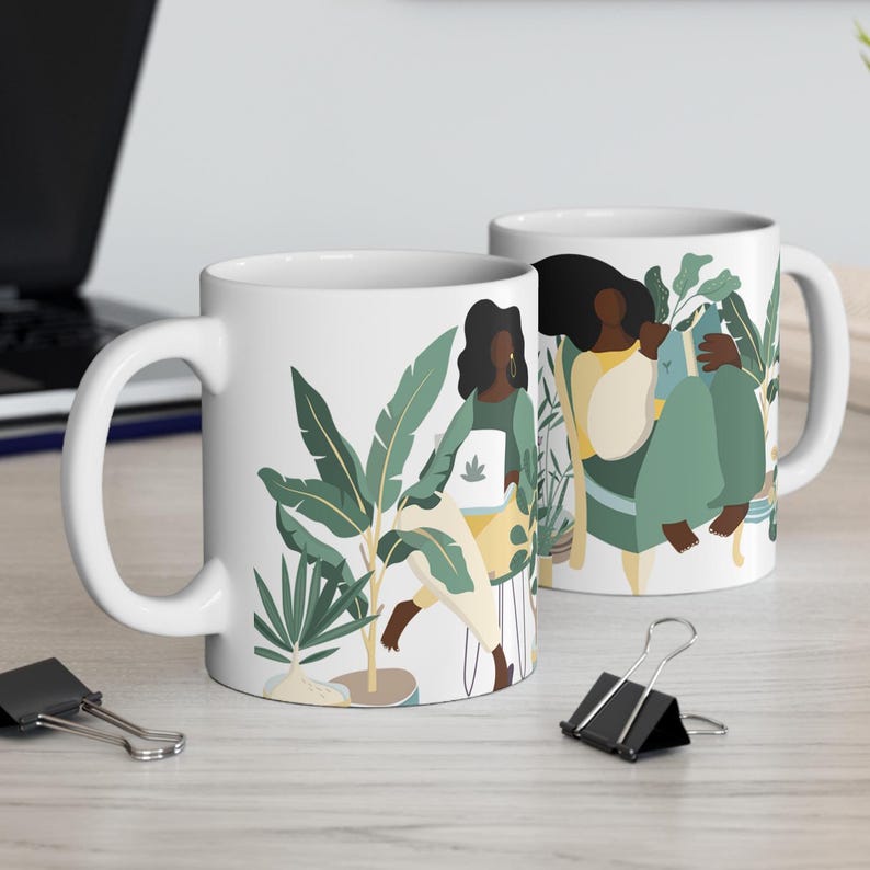 May include: Two white ceramic mugs with a design of two women standing in a jungle setting with plants and flowers. The women are wearing yellow and green clothing and have dark skin and hair.