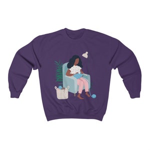 May include: A purple sweatshirt with a graphic of a person sitting in a chair and knitting. The person has dark skin and is wearing a pink dress with white polka dots. There is a basket of yarn next to the chair.