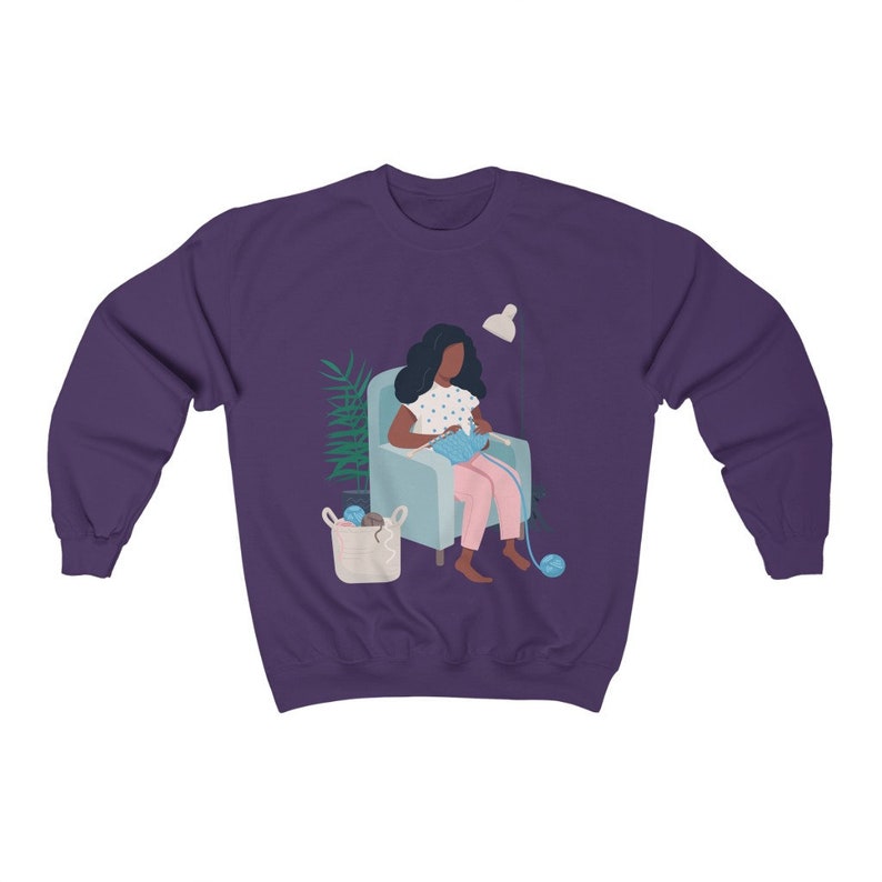May include: A purple sweatshirt with a graphic of a person sitting in a chair and knitting. The person has dark skin and is wearing a pink dress with white polka dots. There is a basket of yarn next to the chair.