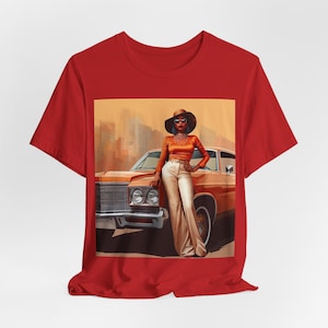 Op de afbeelding: Een rood T-shirt met een grafische print van een vrouw in een bruine hoed en zonnebril die naast een vintage auto staat. De auto is bruin en heeft een wit interieur. De vrouw draagt een bruin topje, een witte broek en een ketting.