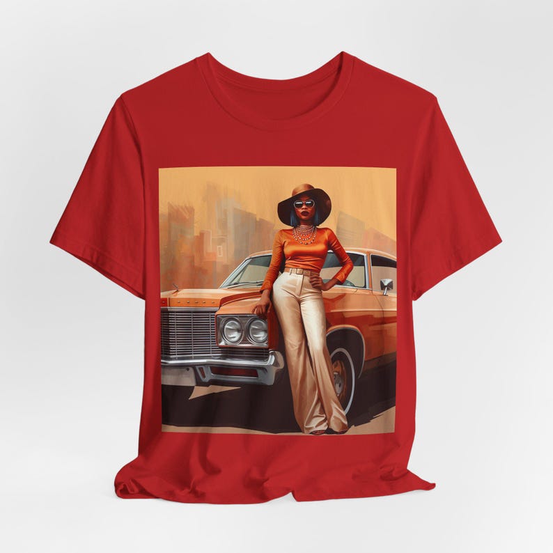 Op de afbeelding: Een rood T-shirt met een grafische print van een vrouw in een bruine hoed en zonnebril die naast een vintage auto staat. De auto is bruin en heeft een wit interieur. De vrouw draagt een bruin topje, een witte broek en een ketting.