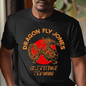 Puede incluir: Camiseta negra con un gráfico naranja de un dragón con el texto "Dragon Fly Jones Self-Defense Training"