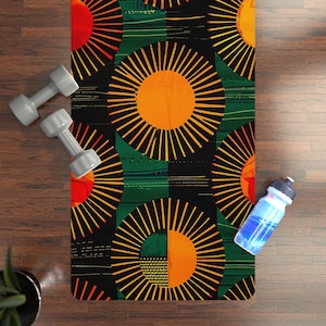 May include: A rectangular yoga mat featuring a repeating pattern of large orange and yellow sunburst motifs on a black and dark green background, accompanied by two light gray dumbbells and a clear plastic water bottle with a blue liquid.