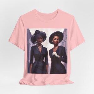 May include: A pink t-shirt featuring a graphic print of two women in black dresses and hats. The women are standing next to each other and looking at the camera.