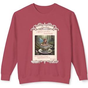 May include: A maroon sweatshirt featuring a framed image of a fairy in a teacup, with the text "Tea Time" at the top. The image includes the quote "Aging is not about losing beauty but gaining a deeper appreciation for it."