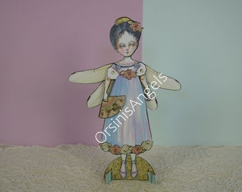 May include: A paper doll with wings, a blue dress with floral accents, and a fan. The doll is standing on a base with the text "OrsinisAngels" printed on it.