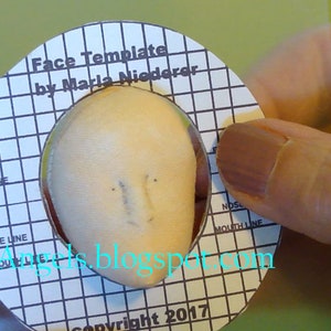 May include: A white face template with a grid pattern and the text "Face Template by Marla Niederer" and "Copyright 2017". The template is being used to draw facial features on a small, round, beige object.
